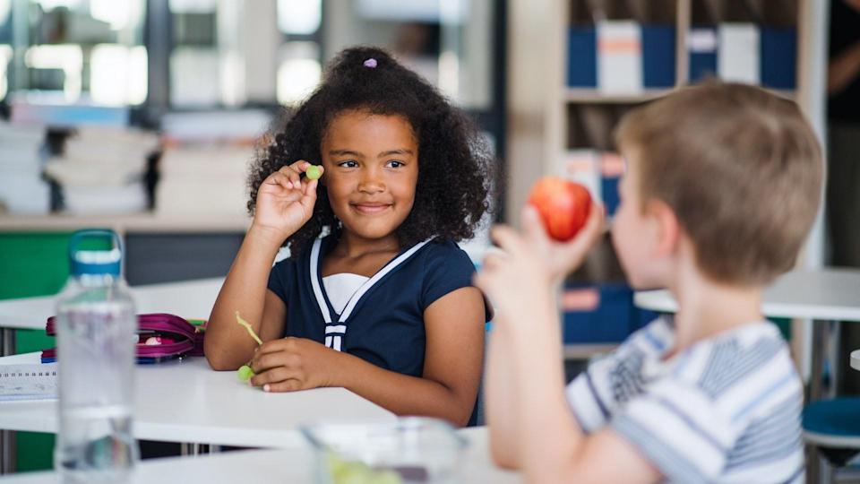 Small school children sitting at a desk in the classroom and eating fruit for a snack.
